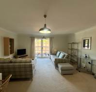 A living room with plaid sofas, a TV, lamp, and a glass shelving unit. A table with chairs is visible. Double doors open to a small balcony with outdoor scenery.