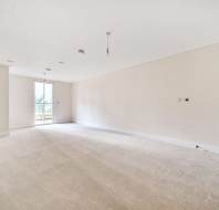 Empty room with beige carpet, white walls, and ceiling. A door with a glass panel leads to a balcony. Light fixtures and electrical outlets are visible.