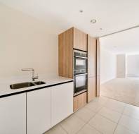 Modern kitchen with white cabinetry, built-in oven, sink, and wooden accents, leading to a carpeted open living area with natural light.