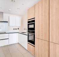 Modern kitchen with white and wood cabinetry, built-in oven, electric cooktop, sink, and white backsplash. Neutral tiled floor and recessed ceiling lights complete the look.