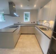 Modern kitchen with beige cabinets, built-in oven, stovetop, extractor hood, and a sink with a window above. Light tiled flooring complements the minimalist design.