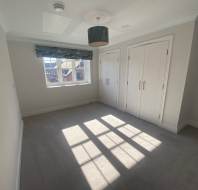 A bright, empty bedroom with a window, light-colored carpet, built-in wardrobes, and a ceiling light fixture. Shadows cast on the floor by sunlight through the window.