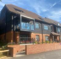 A modern, multi-story building with a brick and wood exterior, featuring a balcony and landscaped front area under a partly cloudy sky.