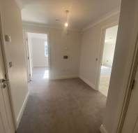 A hallway with beige carpet leads to two open doors revealing sunlit rooms. The walls are painted white, and a single ceiling light is turned on.