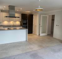 Modern kitchen and living area with white cabinetry, stainless steel appliances, an island, and tiled flooring transitioning to carpeted space. Pendant light hangs above the room.