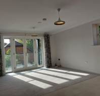 A bright, empty room with two hanging lights, a glass door leading to a small balcony, colorful curtains, and a small window. Sunlight casts shadows on the carpeted floor.