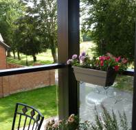 A view from a balcony featuring a hanging planter with pink and purple flowers, a potted plant on a table, and a grassy area with trees and a brick building in the background.