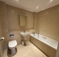 Modern bathroom with beige tiles, featuring a bathtub, sink, toilet, wall mirror, toilet paper holder, and waste bin.