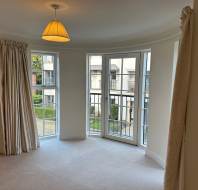 A room with a bay window, cream curtains, and beige carpet. The view includes a garden and building exterior. A ceiling light with a lampshade is turned on.