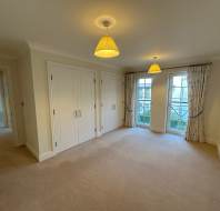 A carpeted empty room with two ceiling lights, a double closet, and French doors leading to a balcony. Cream walls and curtains complement the neutral decor. Open doorway to another room.