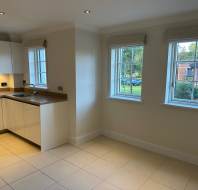Bright kitchen with white cabinets, a small sink, and two large windows. The room has cream tiled floors and a view of greenery outside.