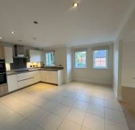 A modern kitchen with white cabinets, built-in appliances, and tiled flooring. Large windows let in natural light. Adjacent dining area with wooden flooring is visible.