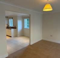A room with beige carpet and a yellow lamp, adjoining a tiled kitchen area with white cabinets and a window.