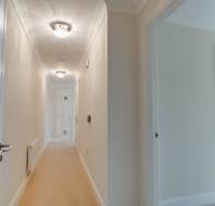 A long, carpeted hallway with white walls and ceiling, illuminated by ceiling lights. It leads to a closed door, with an open doorway visible on the right side.