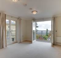 Empty room with beige carpet, two French doors leading to balconies, and two ceiling lights. Walls are cream-colored, and radiators are beneath the windows. A scenic view is visible outside.