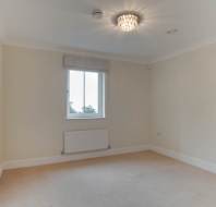 Empty beige room with a window, light fixture on the ceiling, and a radiator below the window. Carpeted floor and white door slightly ajar.
