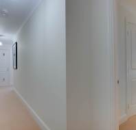A hallway with carpeted floors leads to an open door revealing part of a bedroom with a bed, lamp, and window. Walls are adorned with minimalist artwork.
