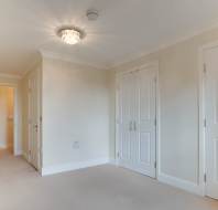 Empty room with cream-colored walls and carpet. Two closed white doors, one open door leading to a hallway. Ceiling light fixture is on.