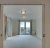 A spacious, empty room with light-colored walls, two ceiling lights, and large double doors leading to a balcony with a view outside.