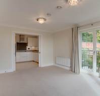 A bright, empty living room with beige carpet, two ceiling lights, and an open view into a modern kitchen. Glass doors lead to a balcony with greenery visible outside.
