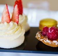 A close-up of assorted desserts on a black tray, featuring pavlova with strawberries, pastries with raspberries, and a yellow macaron.