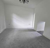 Empty room with gray carpet, white walls, a window on the left, and a black chandelier hanging from the ceiling. A small alcove is visible on the right wall.