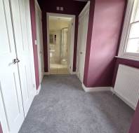 A hallway with purple walls, gray carpet, three white doors, and a window on the right. The view leads to a bathroom with a glass shower enclosure.