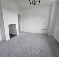 An unfurnished room with light gray carpet, a white fireplace, and a black chandelier. There is a window and a radiator on the right wall.