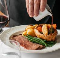 A hand pours sauce over a plate with roast beef, green beans, Yorkshire pudding, potatoes, and vegetables. A glass of red wine is on the table.