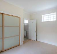 Empty room with cream walls, beige carpet, and a sliding closet door. A white door is open, leading to a tiled bathroom. A small window with grid patterns is above the door.
