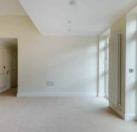 Empty room with beige carpet, white walls, large windows, and a door leading outside. A radiator is visible on the wall near the windows.