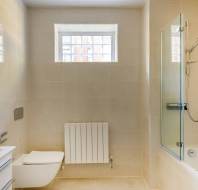 A modern bathroom with a wall-mounted toilet, a sink cabinet, a radiator, and a bathtub with a glass shower screen under a window. The walls and floor are tiled in beige.