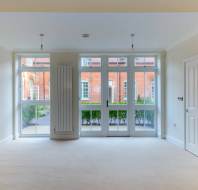 Empty room with light-colored walls and carpet, featuring large windows and a glass door facing an exterior brick building and bushes.