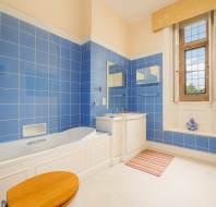 A bathroom with blue tiled walls, a white bathtub, a wooden toilet seat, a vanity with a mirror, a window with patterned glass, and a striped floor mat.