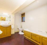 A bathroom with a wood cabinet sink, mirror, toilet, and bathtub. Towels are on a small round table. The room has cream walls and a dark floor. A window has a partially closed blind.