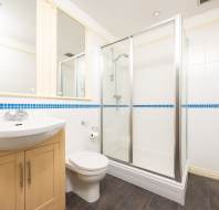 Modern bathroom with a glass shower, white sink, wooden cabinet, and toilet. Blue tile accent line on wall. Matte black floor tiles.