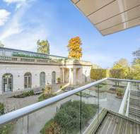 View from a balcony overlooking a historic building with arches, surrounded by a garden and trees under a blue sky.