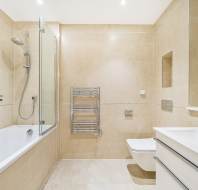 A modern bathroom with beige tiles, a bathtub with a glass shower screen, a wall-mounted toilet, a heated towel rack, and a white vanity with a mirror.