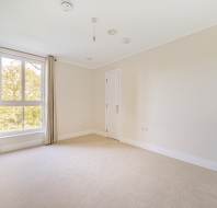 Empty room with beige carpet and cream walls, featuring a large window with curtains, a single door, and ceiling light fixtures.
