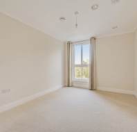 Empty room with cream-colored walls, beige carpet, single window with curtains, ceiling lights, and wall sockets.
