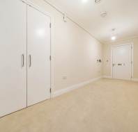 A hallway with beige carpet and cream walls, featuring white double doors on the left and a single white door at the end. Ceiling lights illuminate the space.