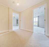 Empty room with beige carpet and walls, featuring two open doorways leading to bright, adjacent rooms. Ceiling lights are on, and windows are visible in the background rooms.