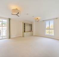 Spacious empty room with large windows, beige carpet, built-in shelving, and ceiling lights. Light streams in from outside greenery.