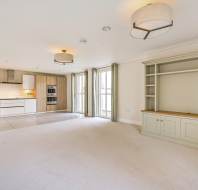 Spacious empty living room with light carpet, built-in shelves, and an open kitchen with wood cabinets. Large windows let in natural light.