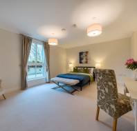 Spacious bedroom with a double bed, large window, armchair, desk, and decorative lighting. Neutral tones and floral accents create a cozy atmosphere.
