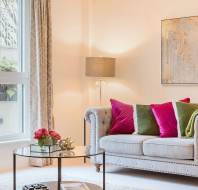 A cozy living room with a beige sofa, pink and green cushions, a green throw, a glass coffee table with a vase of flowers, a lamp, and a window with curtains. A framed abstract art piece on the wall.