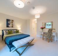 A spacious bedroom with a large bed, blue bedding, and a bench. There is a desk with a mirror and chair, two lamps, and a wall painting. The room is well-lit and has a modern decor.