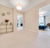 A spacious hallway with cream walls and carpet features two doorways leading to bright rooms, a decorative console table with flowers, and wall decor.