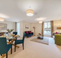 Spacious living and dining area with blue chairs, two couches, a coffee table, and a dining table set. Large windows with curtains let in natural light. Neutral tones and modern decor.