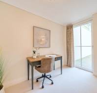 A minimalist study room with a wooden desk, a brown chair, a decorative plant, framed wall art, and a large window with curtains.